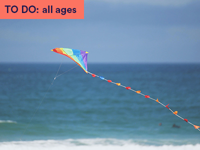 Rainbow kite flying through air across blue sky and sea. Keyword in corner: TO DO: all ages