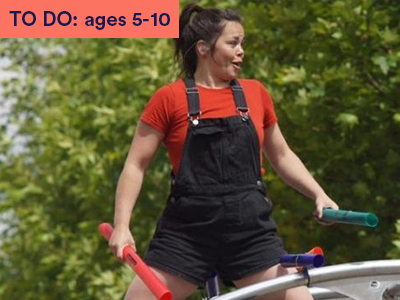 Female dancer wearing red top and black dungarees stands holding instruments. Keyword in the corner TO DO: ages 5-10