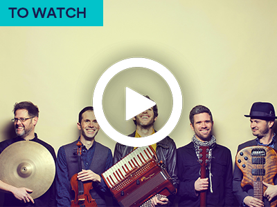 Photograph of MABON band members smiling and holding instruments. White play button overlay. Keyword in the corner TO WATCH