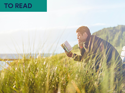 Man sitting on bench outside in green coastline reading a book Keywords: TO READ