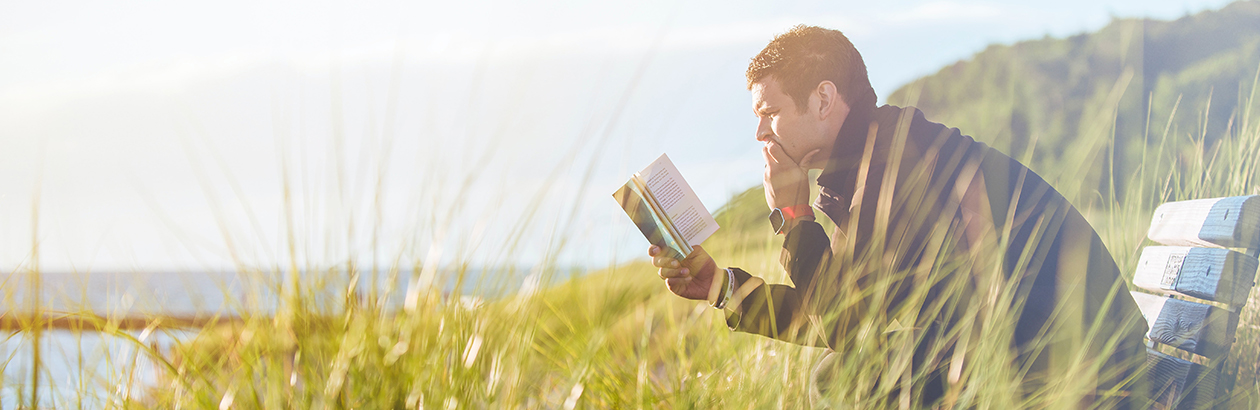 Man sitting outside on bench by coastline reading a book